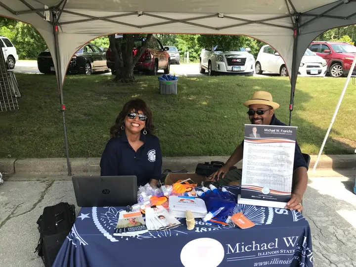 Two vendors under a tent at a table with a laptop and a banner.