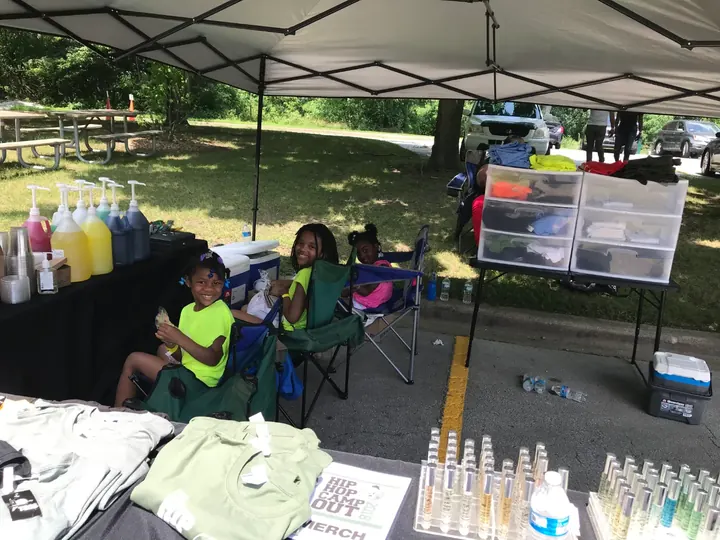 Kids sitting under a vendor tent behind a table of goods.