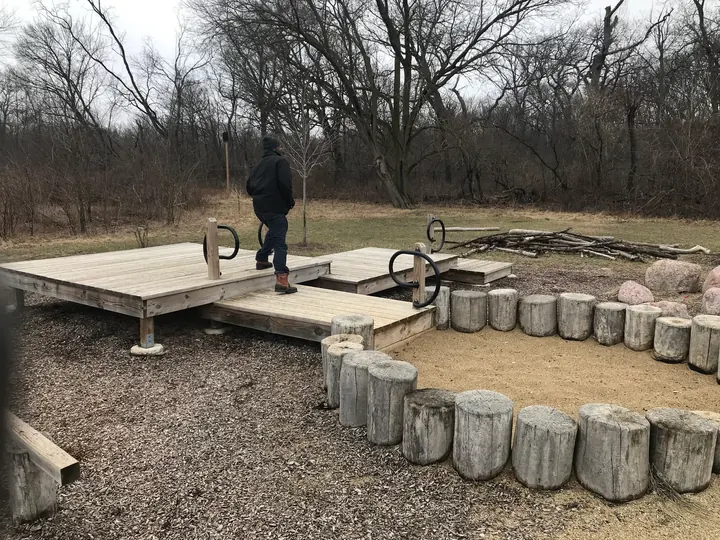 A wooden tent platform and stump seats at a campsite, with a person walking past.