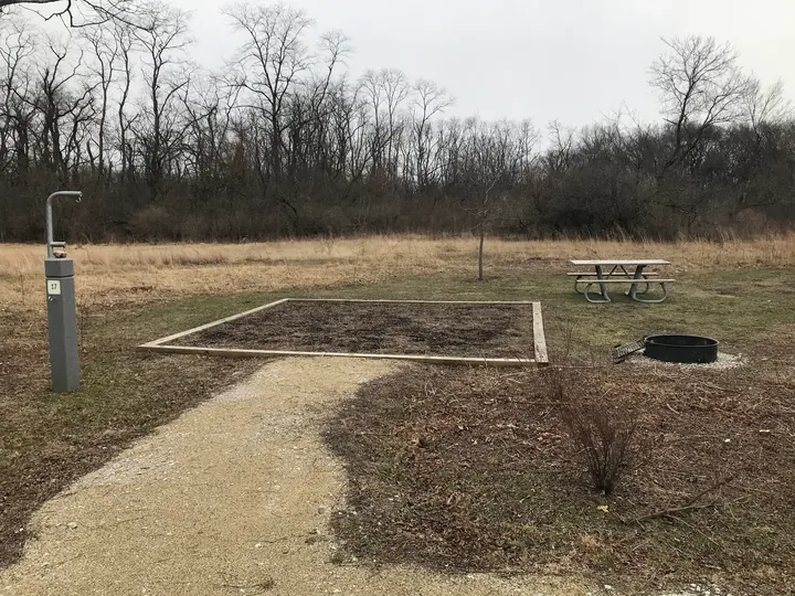 A gravel campsite with a fire pit and a picnic table, edged by trees.