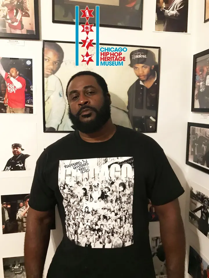 A man in a black graphic T-shirt in front of framed photos.