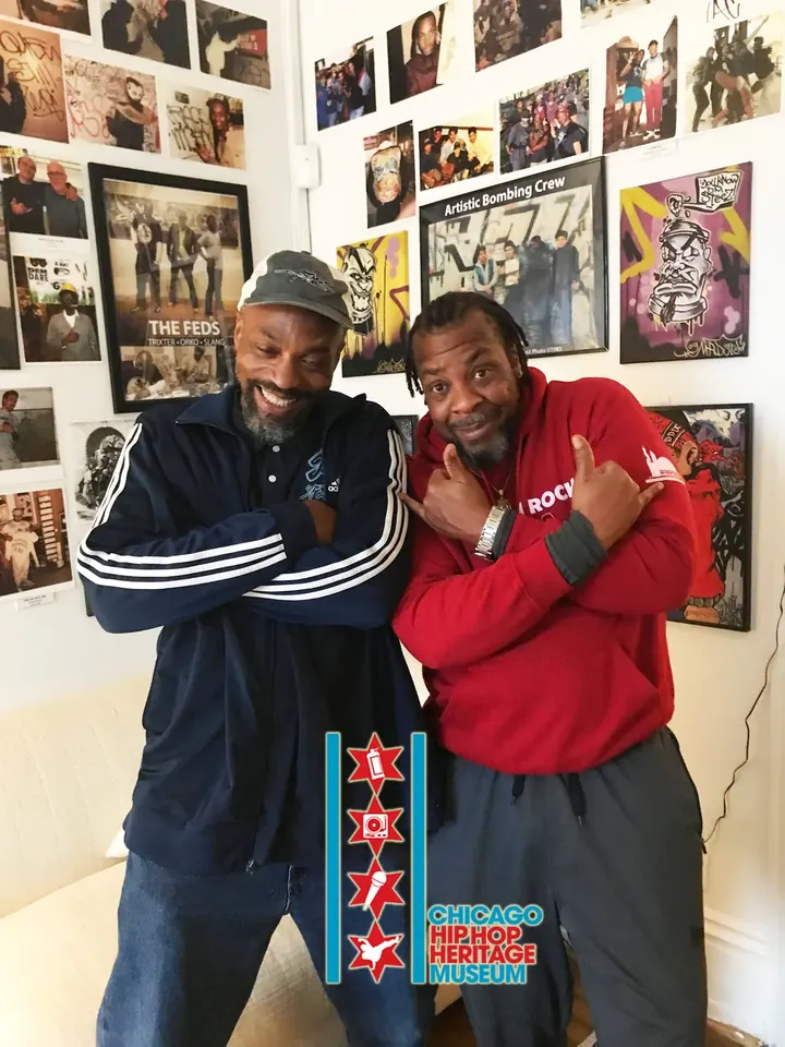 Two visitors hugging in front of the photo wall, one in a red jacket.