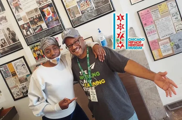 A woman and a man smiling in front of framed photos and flyers.